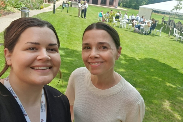 Emma Fromberg and Simona Azzolini stood smiling close together with a group of students sat around round plastic tables in front of a  white marquee