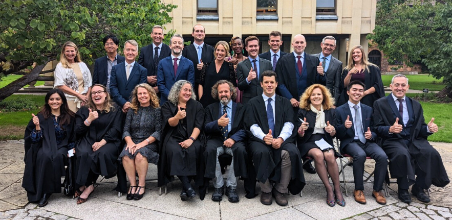 Sustainable Built Environment Graduation Group Photo image of 22 people wearing black formal graduation robes holding their thumbs up and smiling